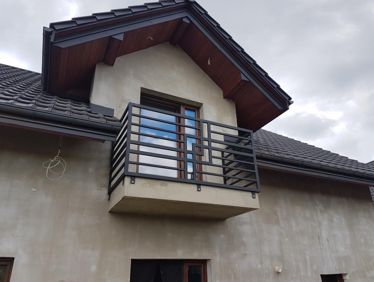 Czarna, spawana balustrada balkonowa z poziomymi prętami, zamontowana na surowym balkonie budynku z szarą elewacją i ciemną dachówką.