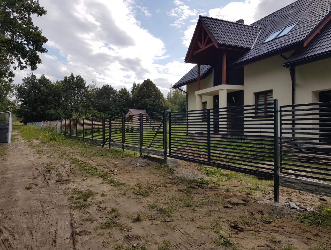 Nowoczesne, poziome, czarne ogrodzenie panelowe z bramą przesuwną na tle domu jednorodzinnego z ciemnym dachem i zielenią; widok z drogi gruntowej.