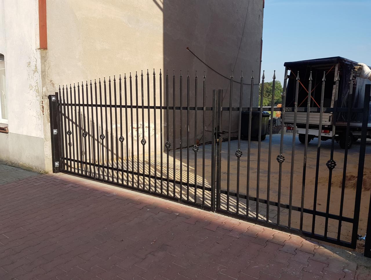 Nowoczesna brama metalowa z pionowymi prętami i ozdobnymi elementami w kształcie rombów, świeżo zamontowana przed budynkiem z kostki brukowej, w tle widoczny samochód dostawczy.