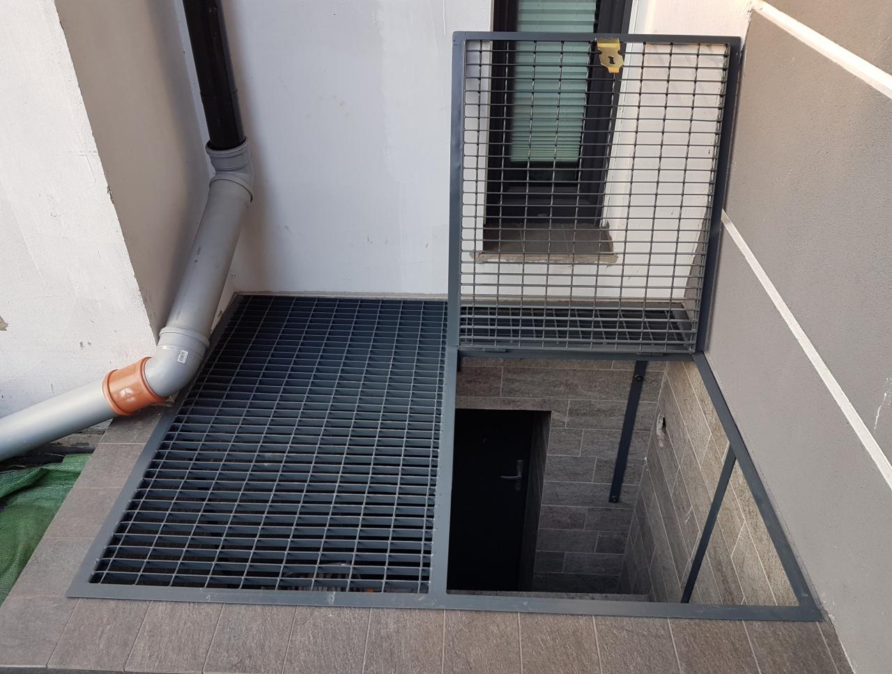 Zabezpieczenie kratą metalową wejścia do piwnicy z uchylną klapą i zamkiem, widoczny fragment rury odpływowej przy ścianie.