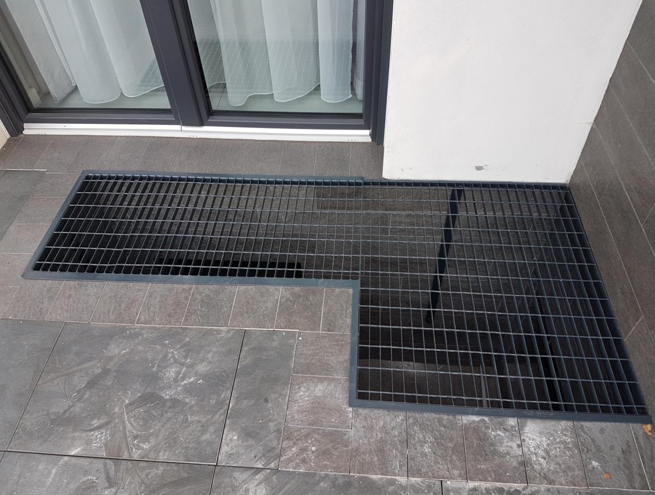 Dwie metalowe kraty pomalowane na szaro, wbudowane w szarą kamienną posadzkę przy ścianie i oknie balkonowym.