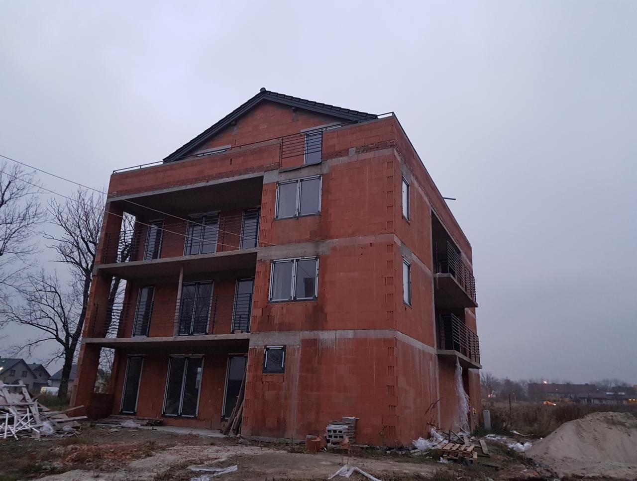 Surowy, trzypiętrowy budynek z czerwonej cegły w trakcie budowy, z widocznymi balkonami z metalowymi barierkami i oknami, na tle pochmurnego nieba.