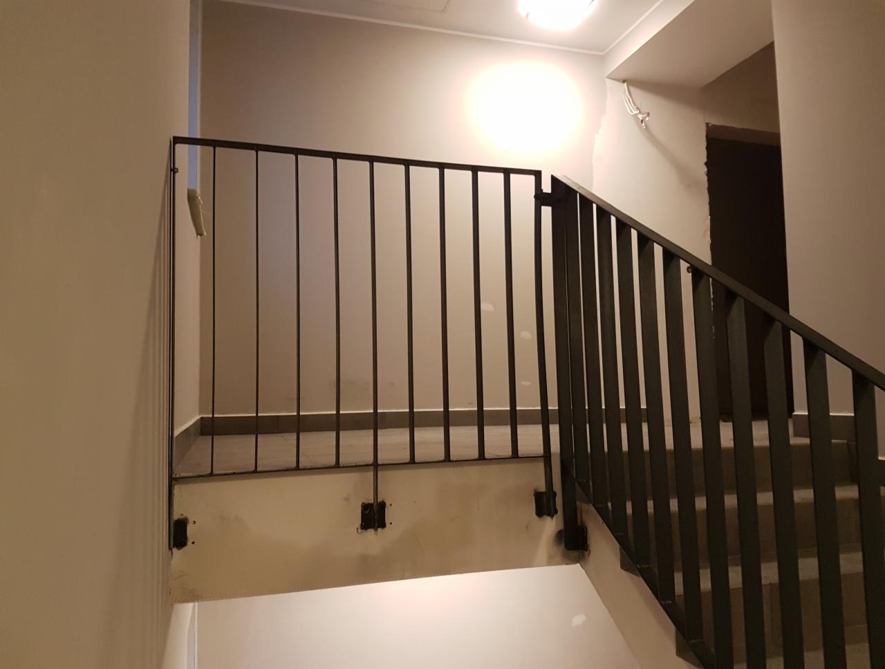 Metalowa balustrada schodowa, czarne pionowe pręty, zamontowana w surowym wnętrzu, widoczne ślady po montażu na ścianie.