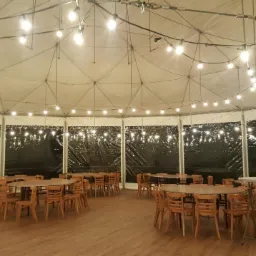 Wnętrze białego namiotu bankietowego z drewnianymi stołami i krzesłami, oświetlonego girlandami żarówek.