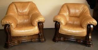 Dwa identyczne, skórzane fotele w odcieniach beżu i brązu, z wyraźnymi przeszyciami i drewnianymi, rzeźbionymi podstawami. Widoczne lekkie ślady użytkowania na siedziskach.