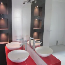 Nowoczesna łazienka z dwoma umywalkami na czerwonym blacie i wnękami z oświetleniem. Minimalistyczny design, białe ściany i płytki podłogowe.