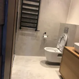 Nowoczesna łazienka z wiszącą toaletą z otwartą deską, grzejnikiem drabinkowym w kolorze czarnym, umywalką nablatową na drewnianej szafce oraz szarymi płytkami na ścianach i podłodze.