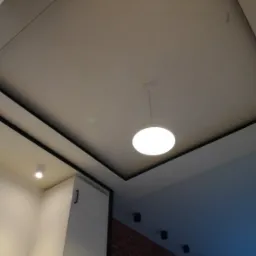 Nowoczesny sufit podwieszany z oświetleniem LED punktowym i centralną lampą wiszącą, czarne obramowania, widoczny fragment białej szafki i ceglanej ściany w tle.