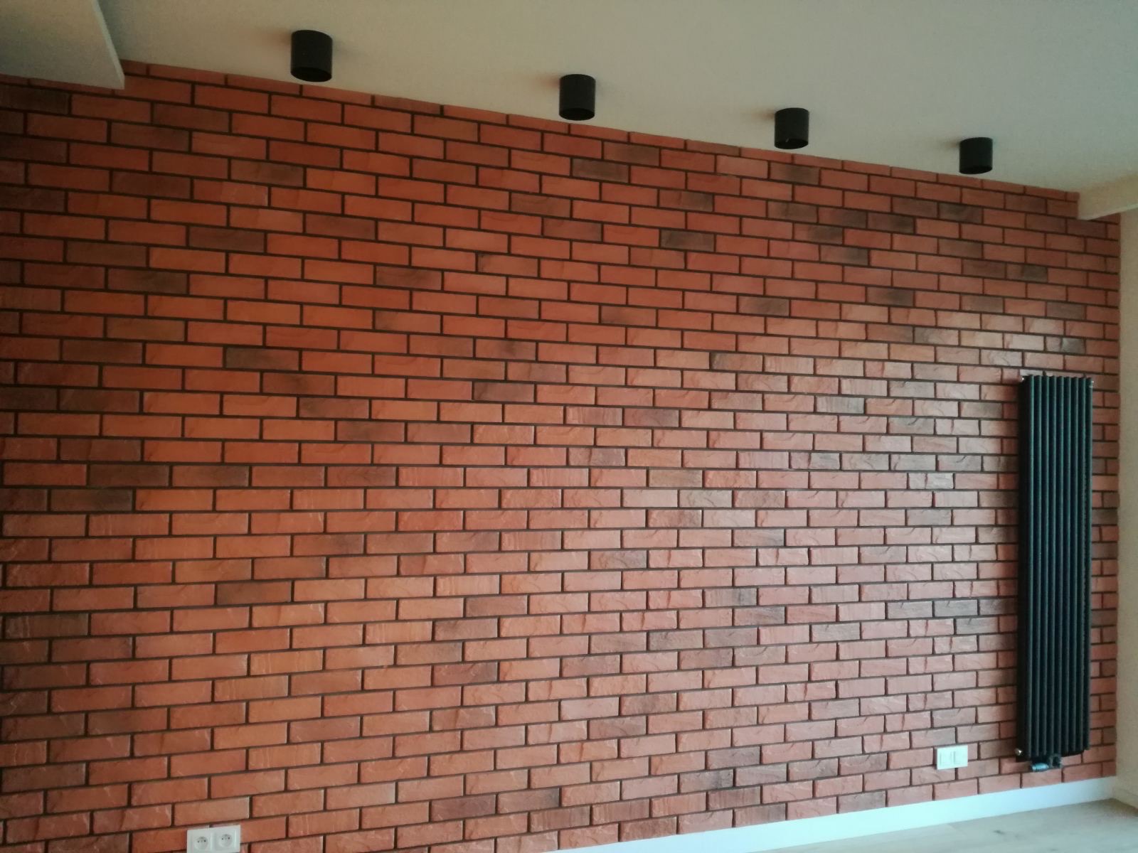 Ściana z cegły klinkierowej w odcieniach czerwieni i brązu, oświetlona czterema czarnymi reflektorami sufitowymi, z czarnym grzejnikiem panelowym po prawej stronie.