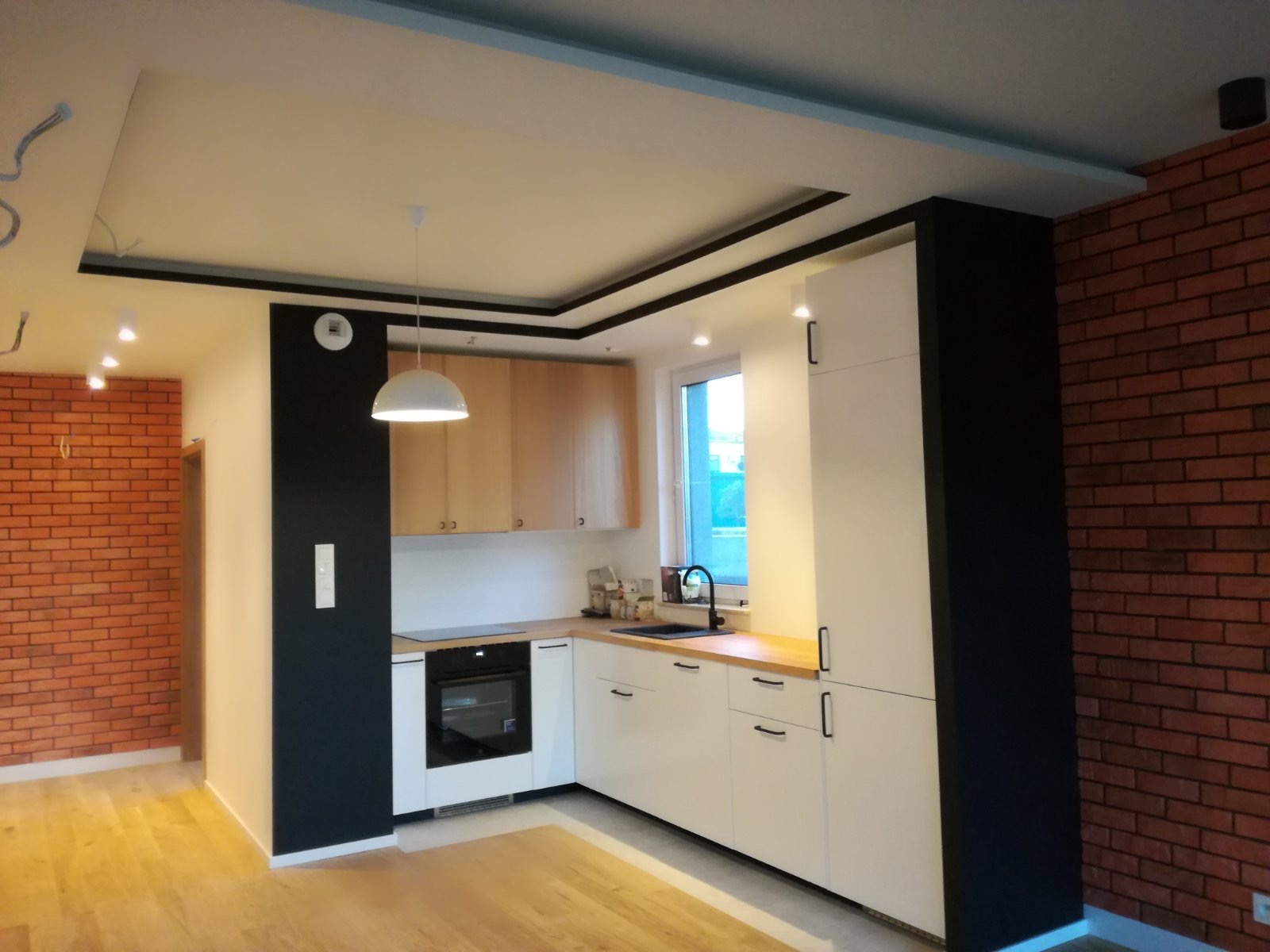 Nowoczesna kuchnia z ceglaną ścianą, białymi szafkami, drewnianym blatem i czarnymi akcentami, oświetlona wiszącą lampą i halogenami.