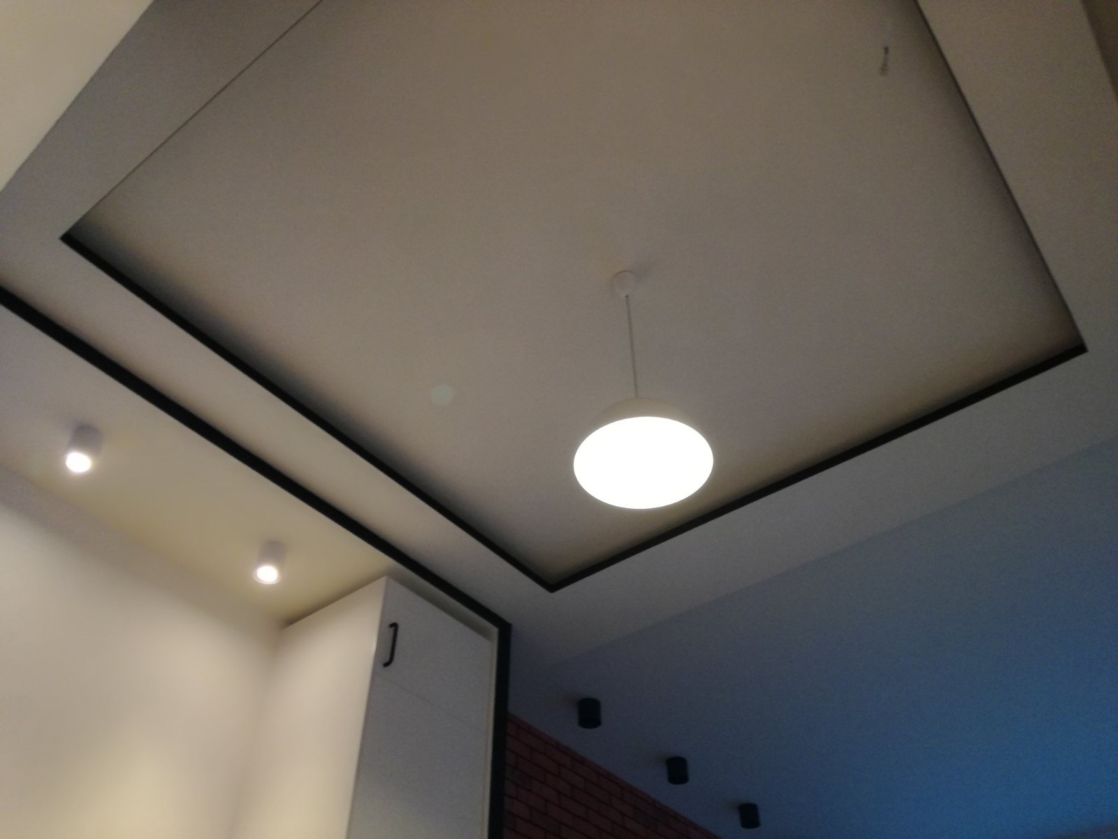 Nowoczesny sufit podwieszany z oświetleniem LED punktowym i centralną lampą wiszącą, czarne obramowania, widoczny fragment białej szafki i ceglanej ściany w tle.