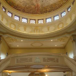 Wnętrze budynku z wysokim sufitem kopułowym, dekoracjami sztukatorskimi i malowidłami, widok z dołu na schody i balkon podczas prac remontowych.