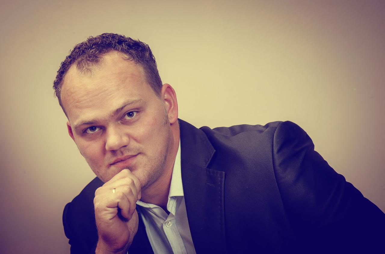 Portret mężczyzny w garniturze, opierającego podbródek na dłoni z widocznym pierścionkiem, na jednolitym tle.