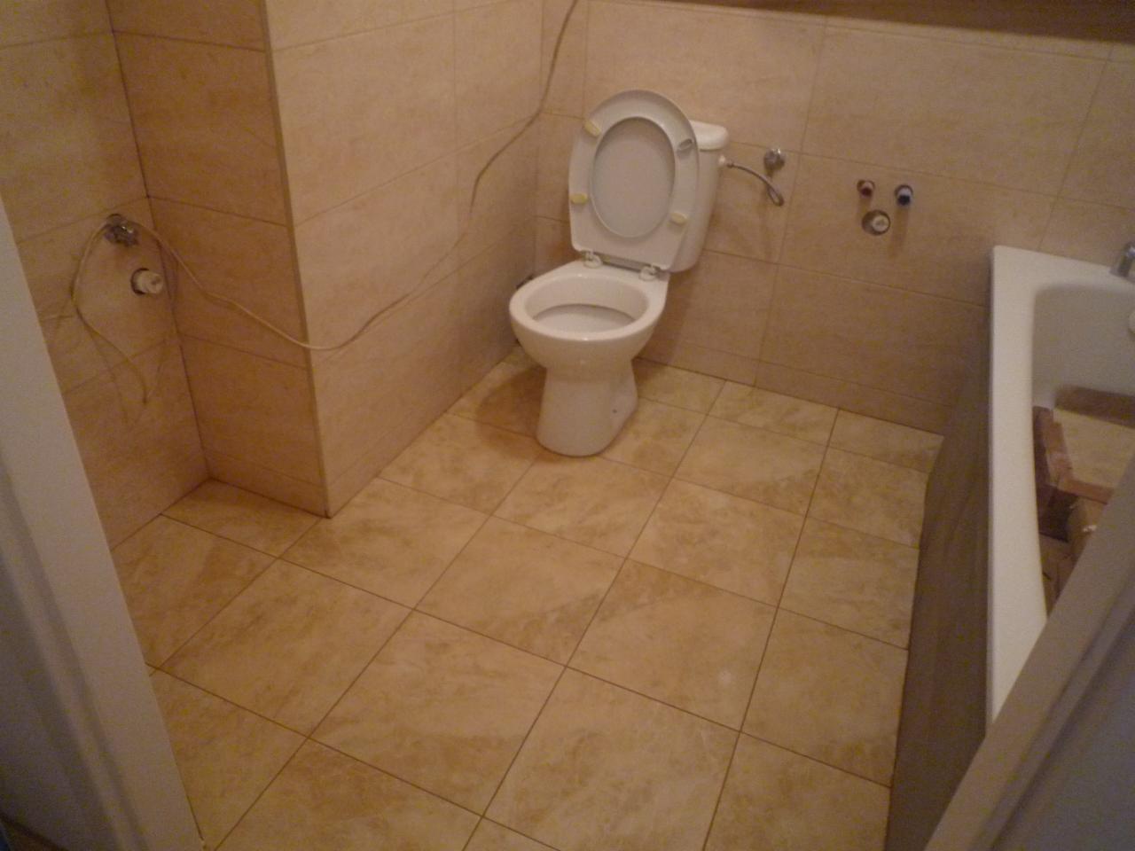 Beżowa łazienka z otwartą klapą toalety, widoczne płytki na podłodze i ścianach, oraz fragment wanny.