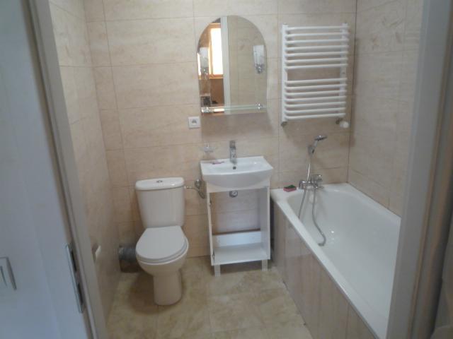 Jasna łazienka z beżowymi płytkami, białą armaturą, grzejnikiem drabinkowym i lustrem z zaokrąglonymi krawędziami, widok z perspektywy drzwi.