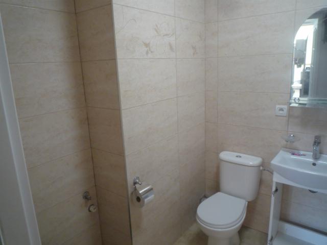 Jasna łazienka z beżowymi płytkami z delikatnym wzorem, widoczna toaleta, umywalka na postumencie i lustro z półką.