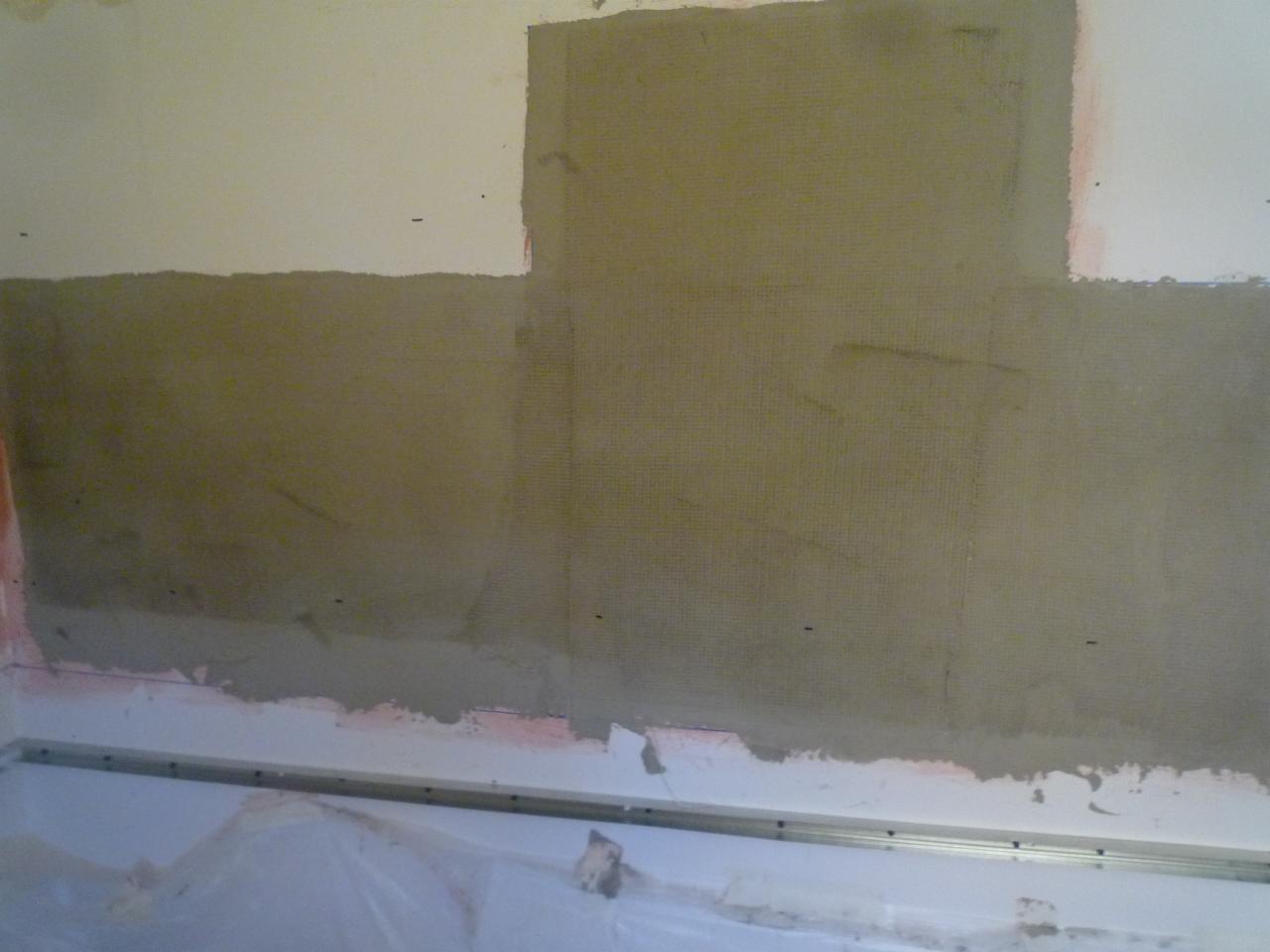 Ściana w trakcie gruntowania i nakładania siatki wzmacniającej pod płytki w łazience, widoczna listwa podłogowa i folia ochronna.