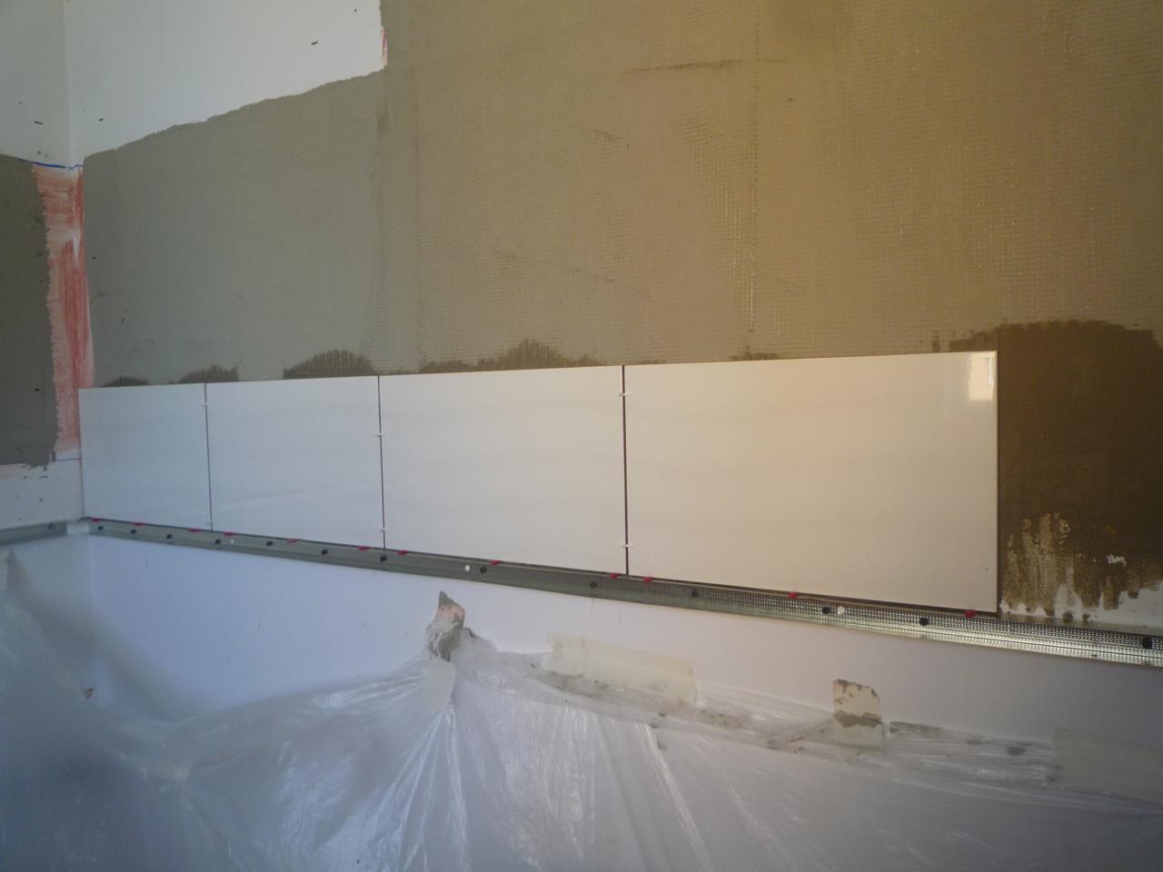 Układanie białych, prostokątnych płytek na ścianie nad wanną, widoczna metalowa listwa poziomująca i folia ochronna na wannie.