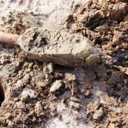 Łopatka z próbką gleby obok wykopanego otworu w piaszczystym podłożu.