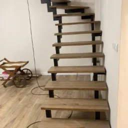 Nowoczesne schody wewnętrzne z drewnianymi stopniami i czarną metalową konstrukcją, widok z dołu, na podłodze widoczny kabel i dekoracyjny wózek.