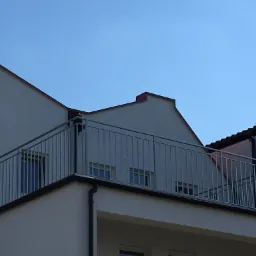 Szara, metalowa balustrada balkonowa z pionowymi prętami na tle jasnego budynku i błękitnego nieba.