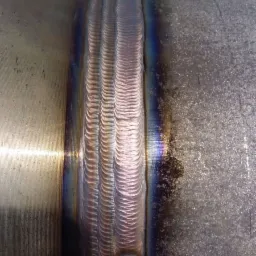 Szczegółowe ujęcie spoiny TIG na rurze ze stali nierdzewnej, widoczne tęczowe przebarwienia termiczne i precyzyjny wzór ściegów.