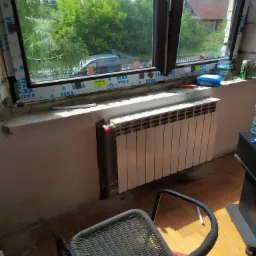 Biały, panelowy grzejnik zamontowany pod oknem z widokiem na zieleń, widoczny fragment krzesła i parapetu z akcesoriami.