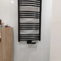 Czarny, łazienkowy grzejnik drabinkowy zamontowany na białej, kafelkowanej ścianie, widoczne zawory termostatyczne, szafka z drewnopodobnymi frontami i akcesoria montażowe w tle.