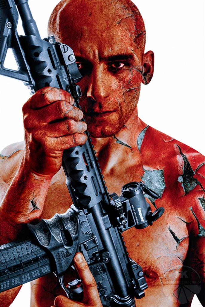 Mężczyzna z czerwoną skórą, imitującą spękanie, trzyma karabin z celownikiem optycznym, patrząc wprost w obiektyw.