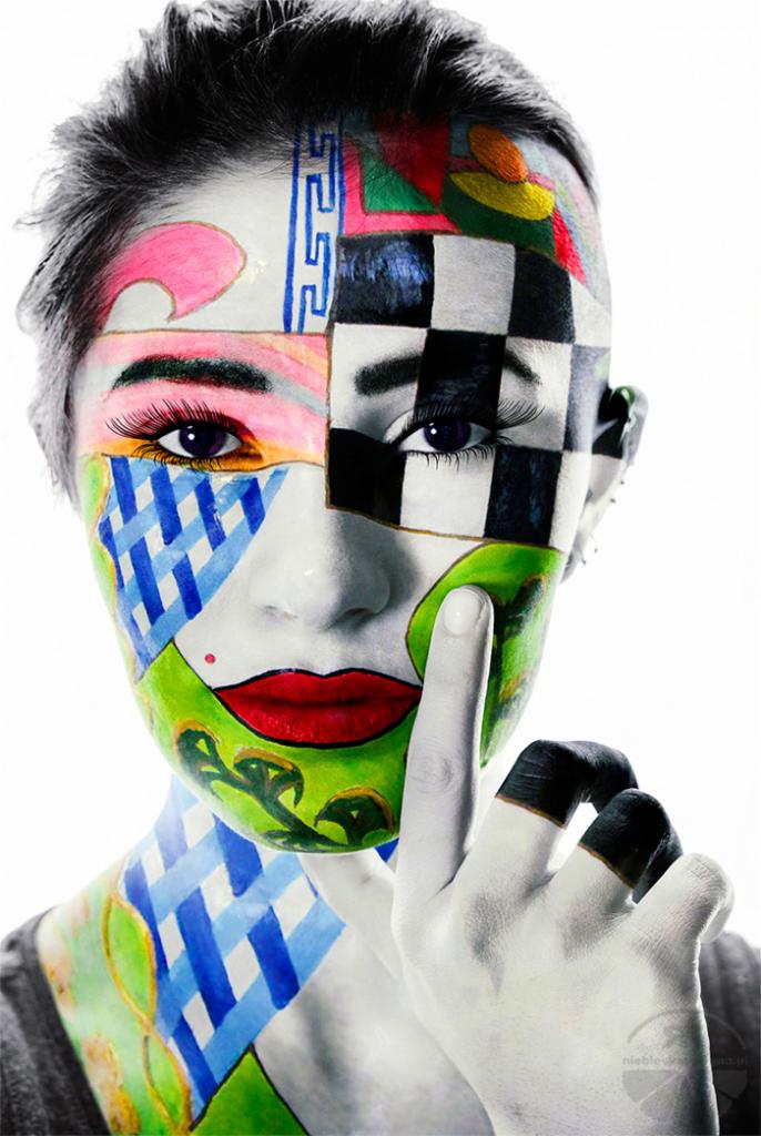 Portret kobiety z artystycznym, geometrycznym makijażem twarzy w odcieniach czerni, bieli, zieleni, niebieskiego i czerwieni, z palcem wskazującym przy ustach w geście uciszenia.