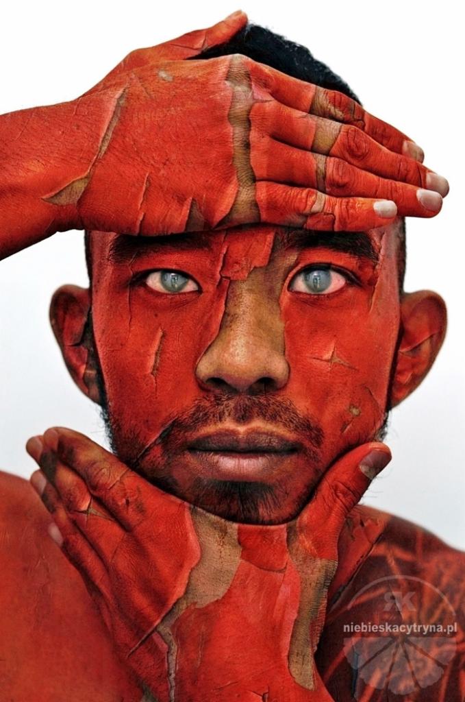 Portret mężczyzny z pomalowaną czerwoną farbą twarzą i dłońmi, imitującą popękaną skórę, z intensywnym spojrzeniem jasnych oczu.