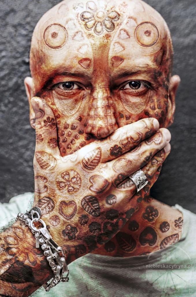 Portret mężczyzny z artystycznymi tatuażami na twarzy i dłoni, zakrywającego usta, z srebrnym pierścieniem i bransoletką