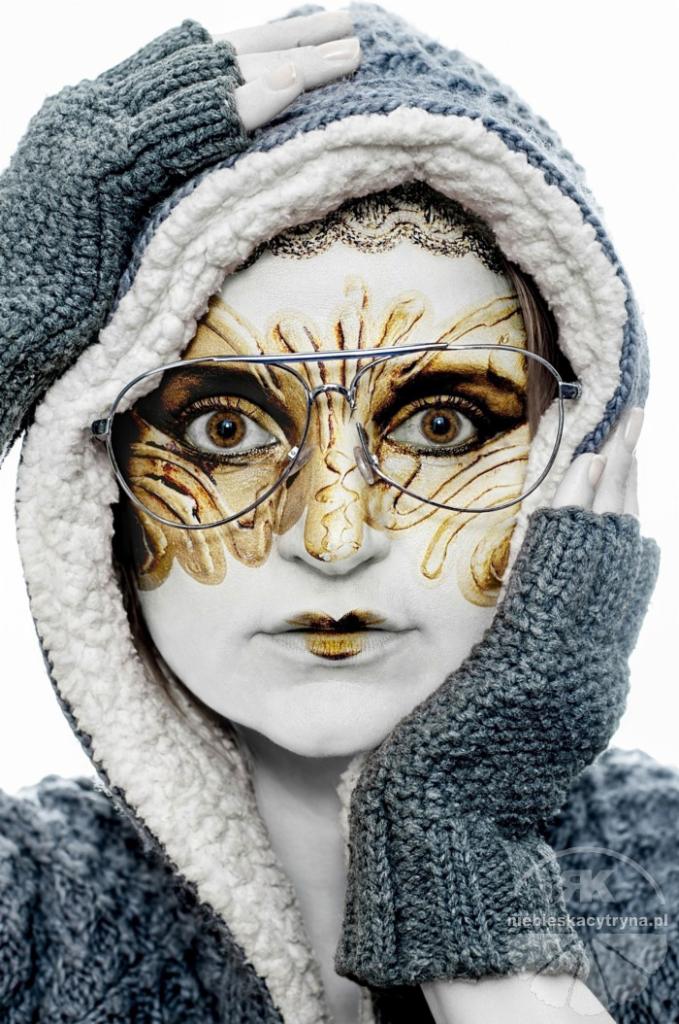 Portret kobiety z artystycznym makijażem twarzy w odcieniach złota i brązu, w okularach typu pilotki i szarym swetrze z kapturem wykończonym białym futerkiem.