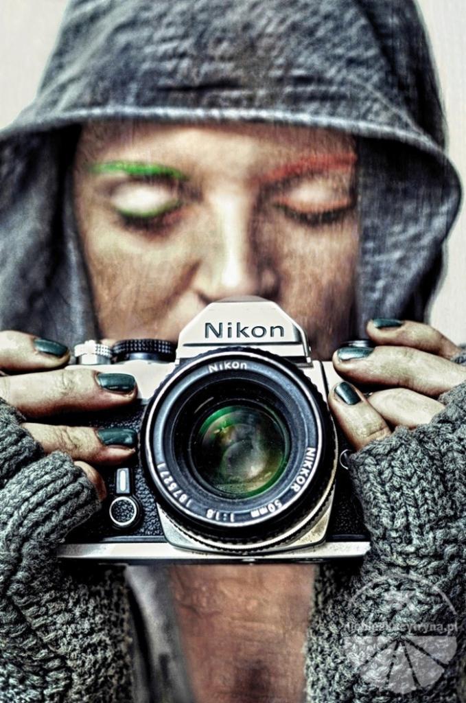 Kobieta w kapturze z zamkniętymi oczami trzymająca stary aparat Nikon z obiektywem 50mm, nietypowy makijaż brwi w kolorach zielonym i czerwonym.