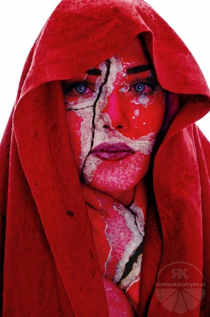 Portret kobiety z intensywnym czerwonym makijażem i pęknięciami imitującymi skorupę na twarzy, ubrana w czerwoną szatę z kapturem, na białym tle.