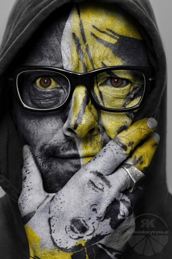 Portret mężczyzny z połową twarzy pomalowaną na żółto i czarno, z okularami i dłonią z namalowaną twarzą przy ustach.