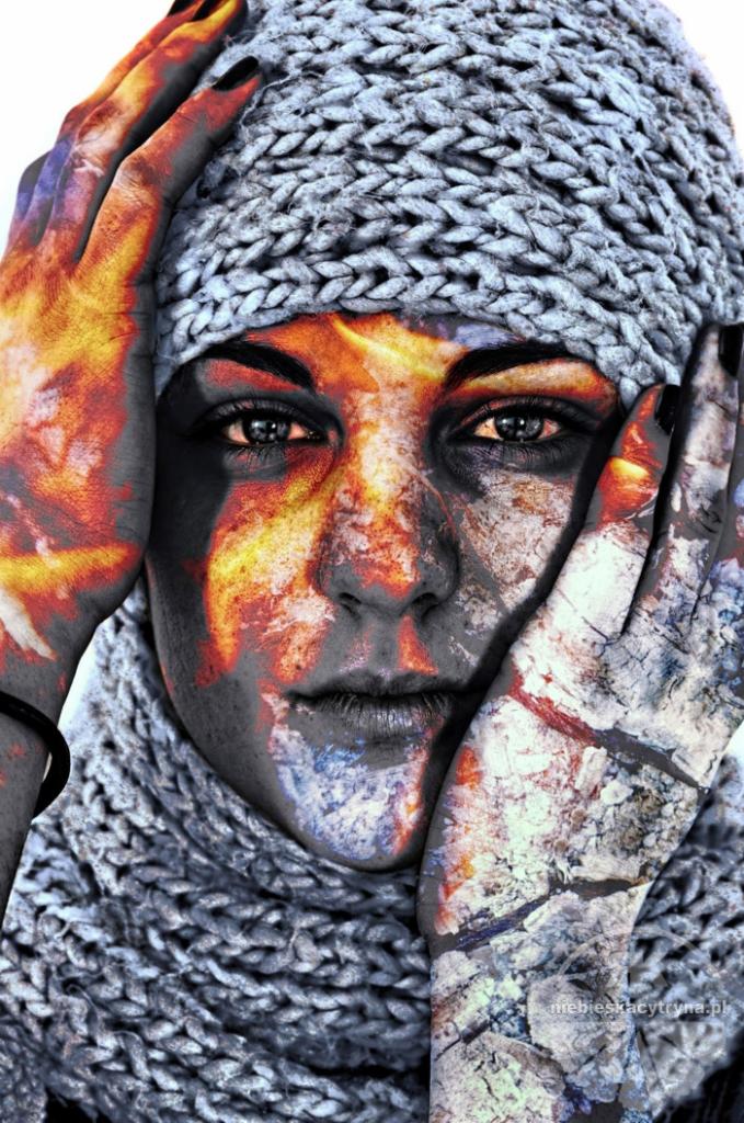 Portret kobiety w wełnianej czapce i szaliku z abstrakcyjnym makijażem w odcieniach ognia i kamienia na twarzy i dłoniach.