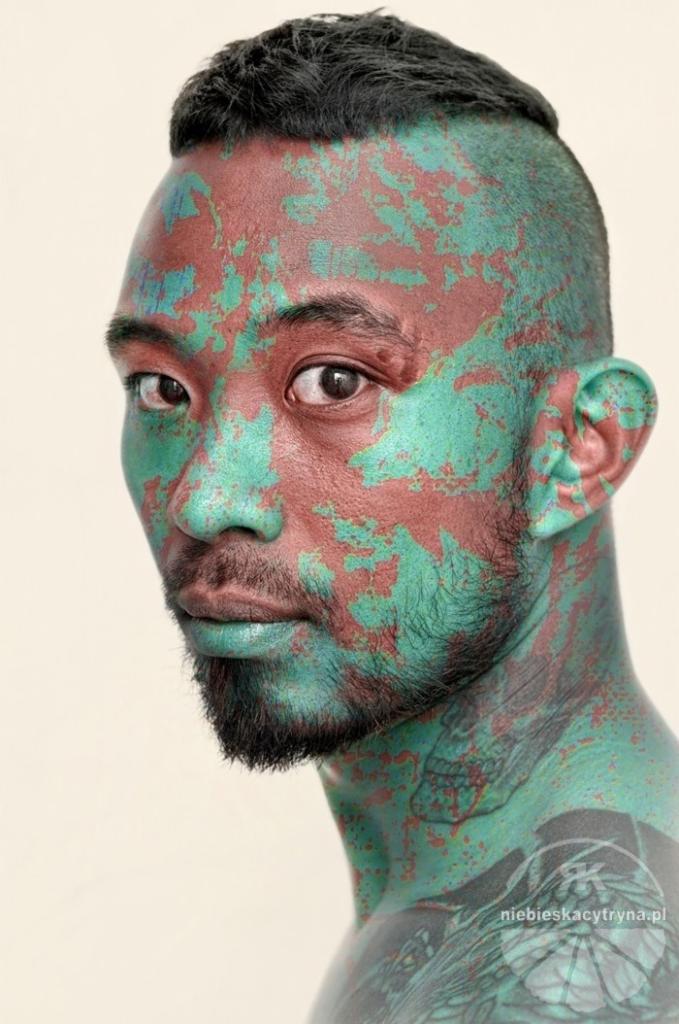 Portret mężczyzny z pomalowaną twarzą i szyją w odcieniach zieleni i brązu, z widocznymi tatuażami na szyi i ramieniu.