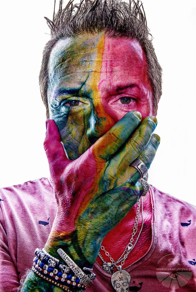 Portret mężczyzny z twarzą i dłonią pomalowaną w tęczowe barwy, zakrywającą usta, z widocznymi srebrnymi pierścieniami i bransoletkami, na białym tle.