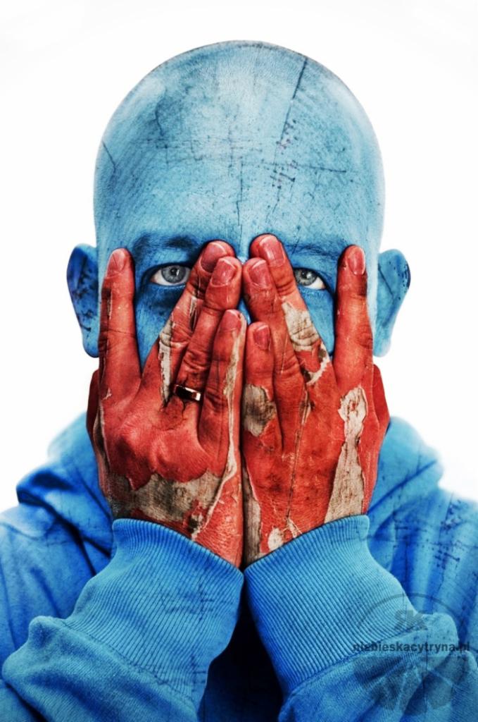 Niebieskoskóry mężczyzna w kapturze zakrywa twarz dłońmi pomalowanymi na czerwono, na palcu złoty pierścionek, intensywne spojrzenie.