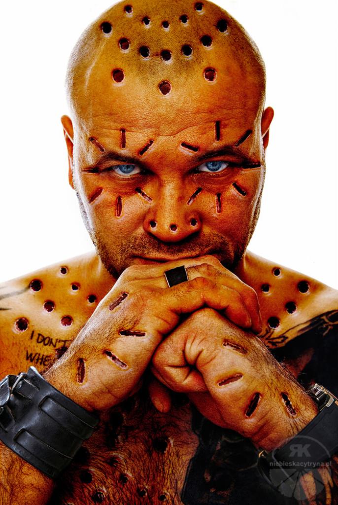 Mężczyzna z intensywnie niebieskimi oczami, licznymi bliznami i otworami na głowie, twarzy i klatce piersiowej, trzymający złączone dłonie z czarnym pierścieniem, na białym tle.