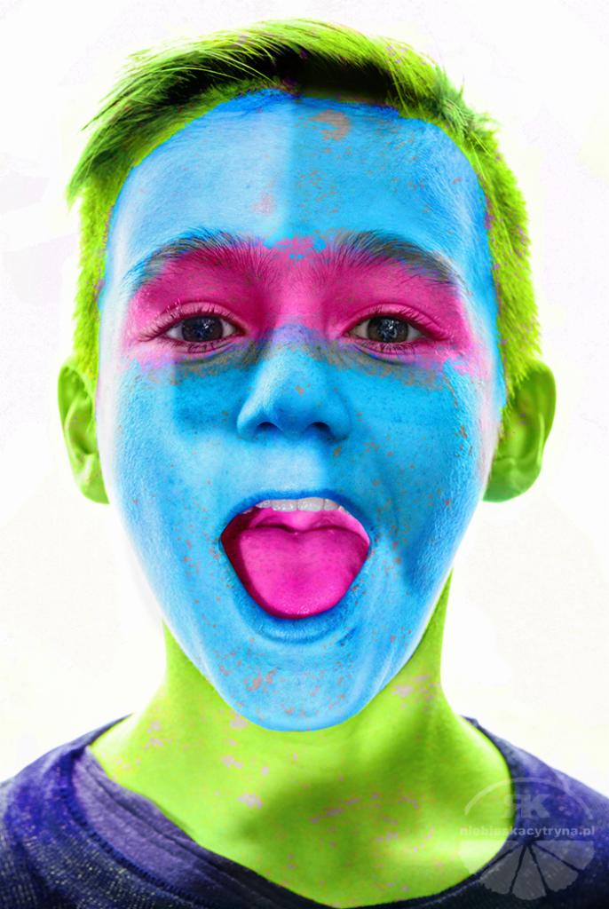 Portret nastolatka z pomalowaną twarzą w odcieniach niebieskiego, różowego i zielonego, otwarte usta z różowym językiem, jasne tło.