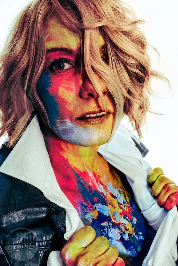 Portret kobiety z artystycznym makijażem w odcieniach niebieskiego, czerwonego i żółtego, imitującym pociągnięcia pędzla na twarzy i ciele, ubrana w białą koszulę i czarną skórzaną kurtkę.