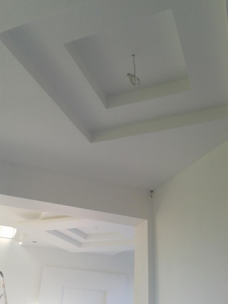 Nowoczesny, biały sufit z dekoracyjnymi wgłębieniami i wystającym okablowaniem w centralnej części. Widoczny fragment drugiego sufitu w tle.