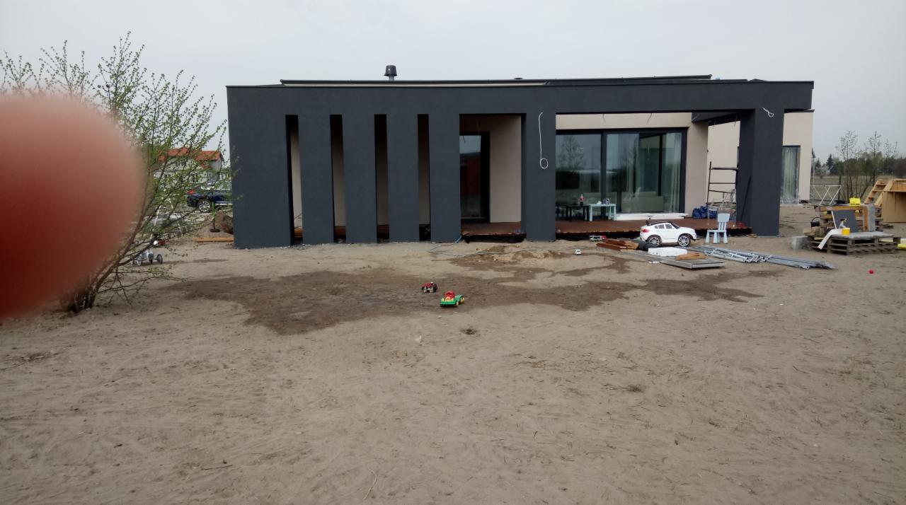 Nowoczesny dom jednorodzinny w trakcie budowy, z ciemnoszarą elewacją i dużymi przeszkleniami, otoczony piaszczystym terenem z zabawkami dziecięcymi i materiałami budowlanymi.