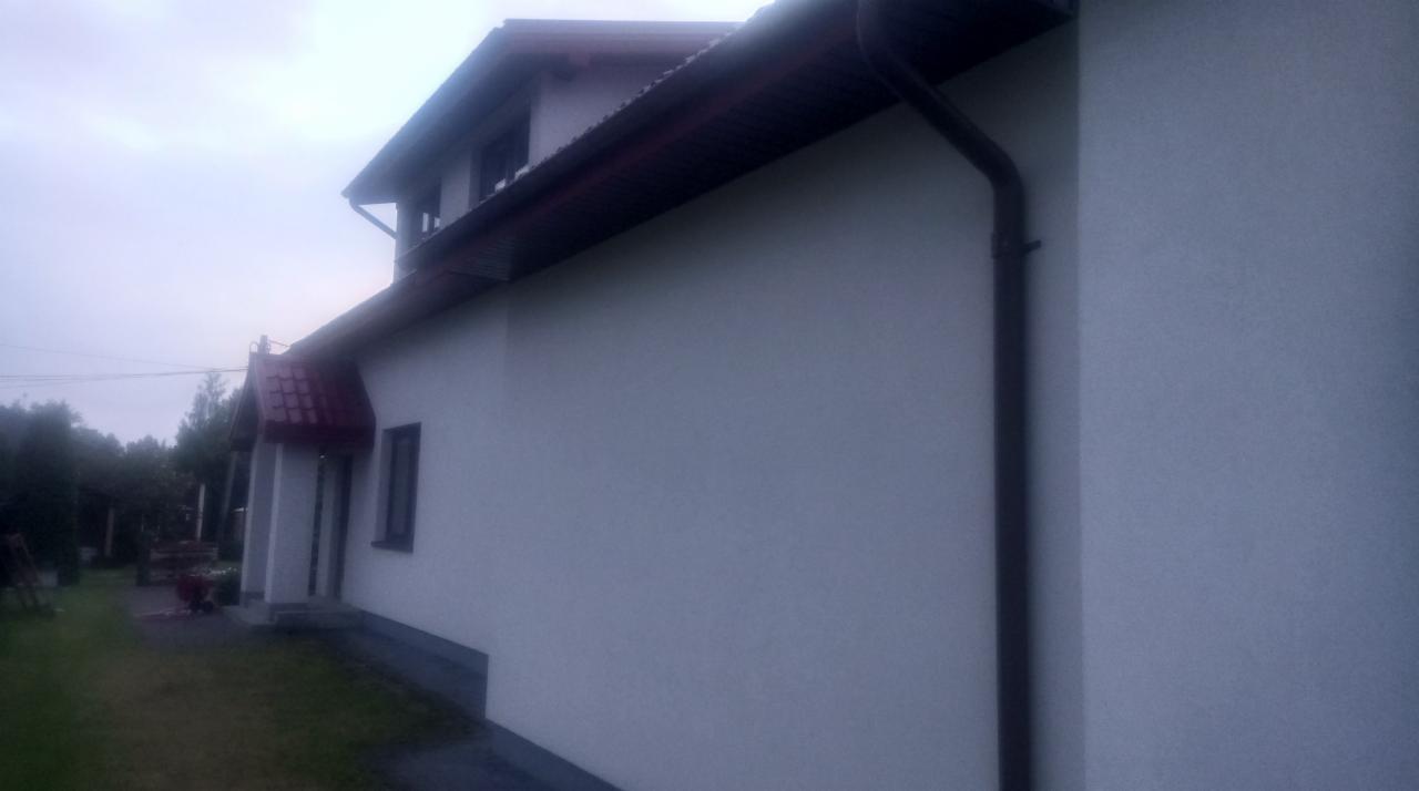 Fragment ściany domu jednorodzinnego z nową, jasną elewacją i brązową rynną spustową, widok z boku w pochmurny dzień.