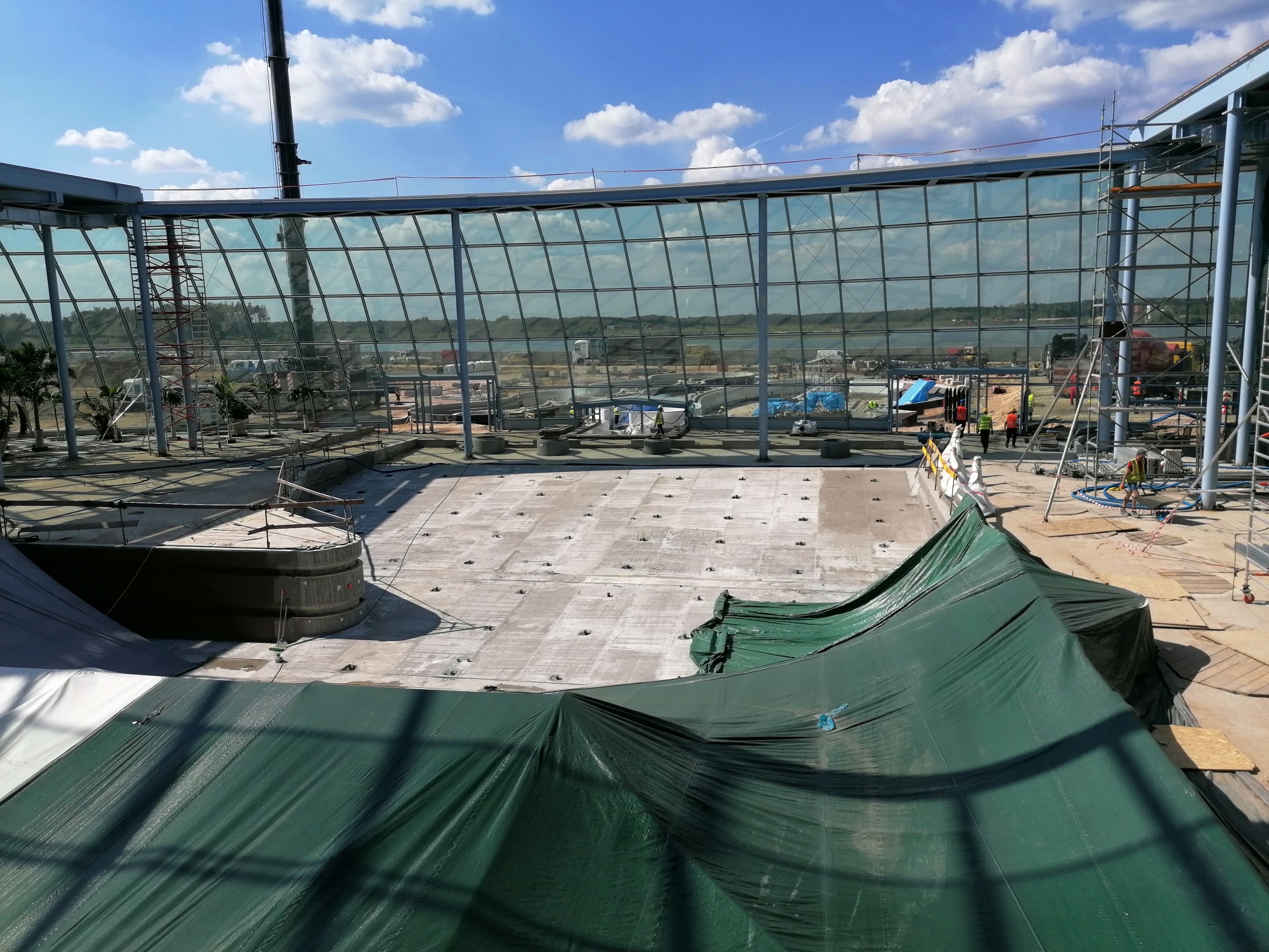 Budowa przeszklonej elewacji budynku w trakcie realizacji, widoczne rusztowania, betonowa podstawa i zielona plandeka ochronna, w tle błękitne niebo z chmurami.