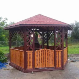 Ośmiokątna konstrukcja z czerwonym dachem, częściowo osłonięta drewnianymi panelami z ażurowymi ścianami, z grillem w środku, usytuowana na betonowym podłożu w ogrodzie.