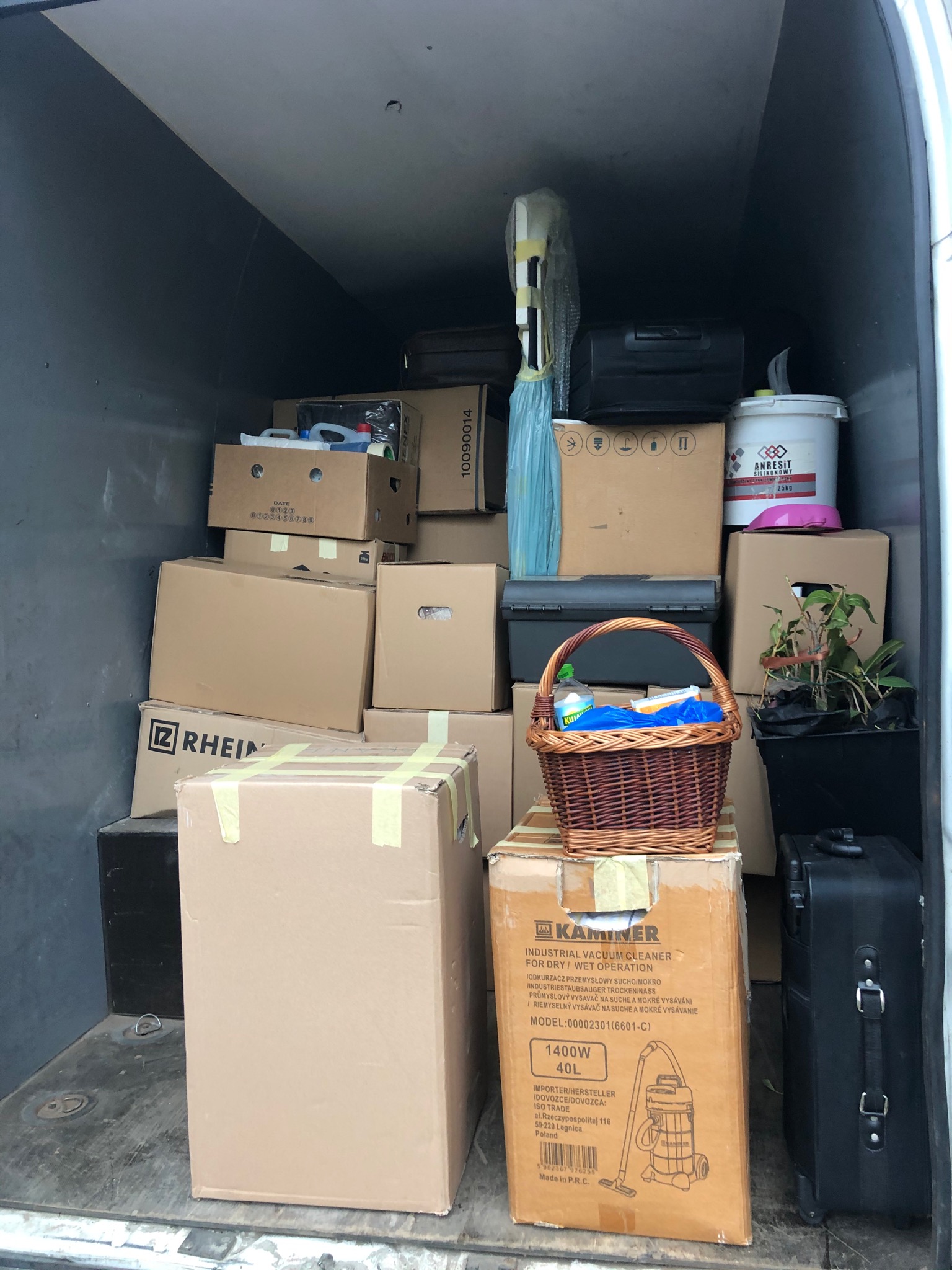 Wnętrze furgonetki wypełnione kartonami, walizką, koszem wiklinowym z niebieską folią i rośliną doniczkową, przygotowane do transportu.