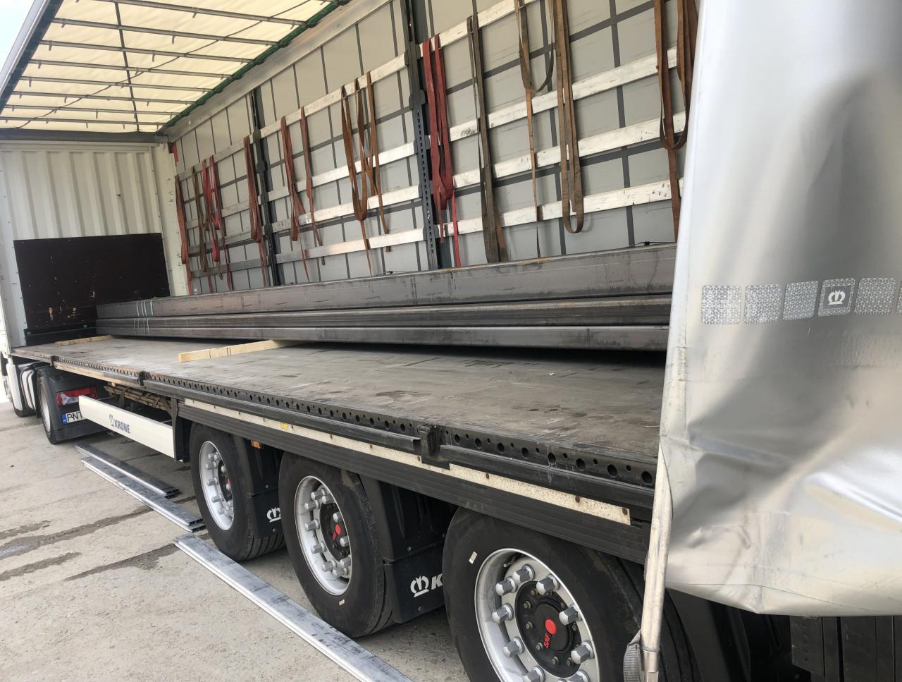 Ciężarówka z naczepą typu laweta przewożąca metalowe profile, zabezpieczone pasami transportowymi, widok z boku na koła i część naczepy.